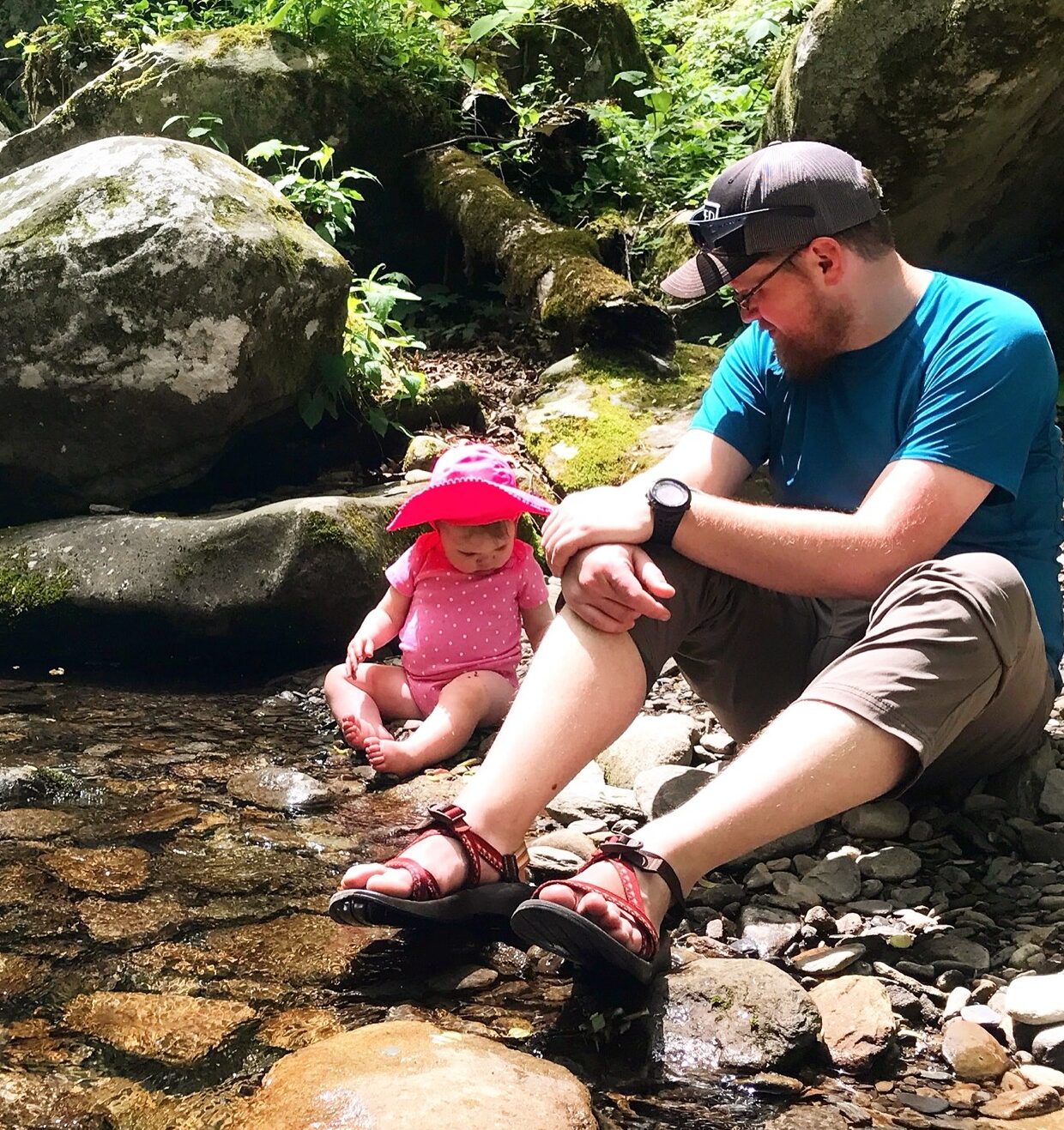 A man and a baby sitting on the side of a brook/river.